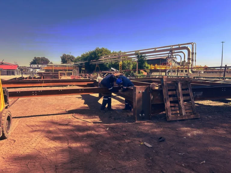Heavy-duty steel beams stacked at a mineral processing site with industrial piping in the background.